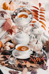 A cup of hot tea with lemon in an early autumn morning by the window. A stack of sweaters, yellow leaves, pumpkin, candles. Decor. Autumn concept of warm drink on a cold day.