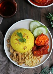 Top view of Nasi Kuning, Indonesian popular traditional  yellow rice served with chicken and eggs balado, noodle,  tempeh, and cucumber slices. Dark wooden background