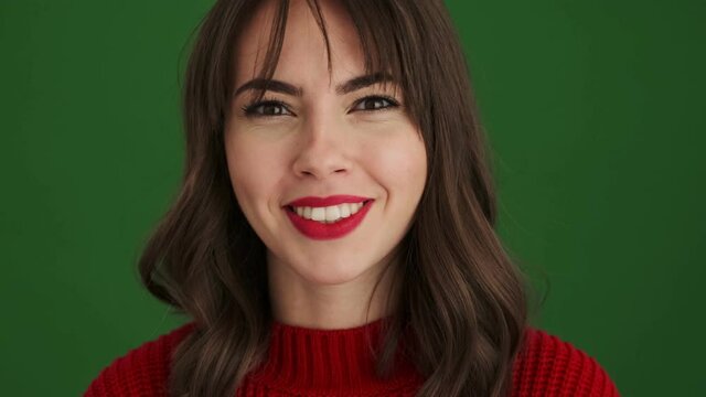 A close-up view of a cheerful woman in a red sweater is showing funny facial expression to the camera standing isolated over green background