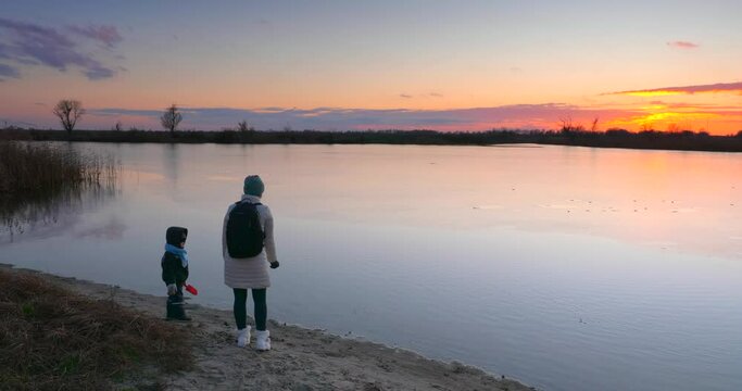 Mother with Child Throw Sand in Water near Wooden Fishing Pier on River Lake Pond Shore. Scenic Picturesque Sunset Cloudscape Reflection Beautiful Evening Landscape