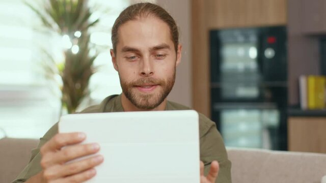 Handsome nice caucasian man using tablet computer tapping on screen scrolling surfing web-pages videochatting texsting messages in the living room. Male face p-ortrait. Devices.