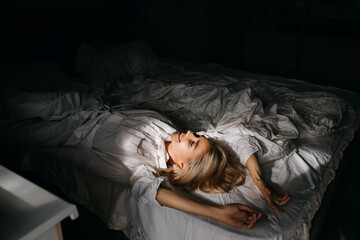 Young blonde woman lying on bed in morning light, smiling.