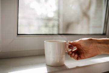 Woman hand holding a cup of coffee with steam by the window.