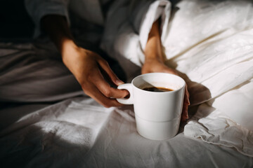 Woman holding a cup of coffee, sitting on bed in the morning light.