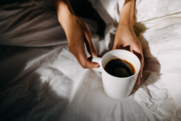 Woman holding a cup of coffee, sitting on bed in the morning light.