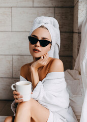 Woman in white bathrobe, a towel on head and sunglasses with a cup of coffee.