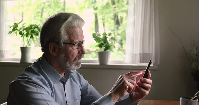 Grey-haired senior man sit at table holds mobile phone text message, writes or send sms. Older generation male use modern gadget chatting in social network, shopping online, spend time with smartphone