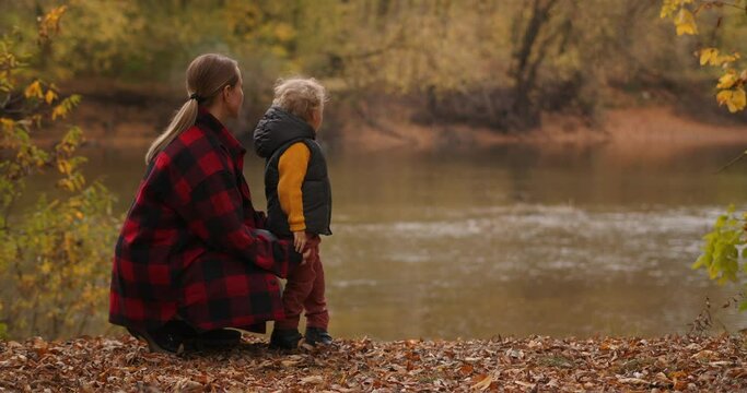 family rest in autumn forest, mother and little son are viewing lake, standing on coast with dry grass and foliage