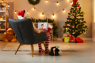 Rear view of Santa Claus sitting in a chair with a laptop and talking on a video call with a child.
