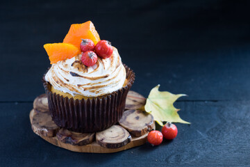 Homemade pumpkin muffin cupcake decorated with meringue, berries and pieces of pumpkin on black wooden table with autumn leaves in background. Selective focus