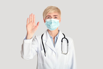 Middle aged female doctor in white coat with stethoscope in protective mask, shows stop sign with hand