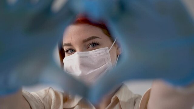 Portrait of woman with heart shaped hands winking and wearing medical face mask and gloves, looking at camera. Close-up. Frontal view