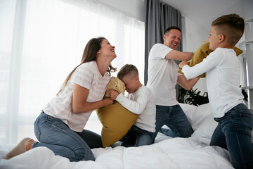 Young parents with sons fighting pillows on the bed. Happy mother and father with boys having fun...