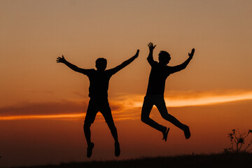 Silhouette of two Indian friends jumping with arms raised against the sky during the sunset	