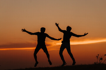 Fototapeta premium Silhouette of two Indian friends jumping with arms raised against the sky during the sunset 