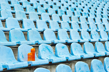 Naklejka premium Lots of empty seats in the stadium. Orange water bottle on the seat. Drink for athletes. Plastic seats are arranged in a row. Dirty and scratched blue fan seats.