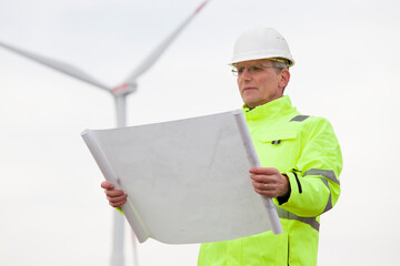 Mature engineer standing in front of a wind turbine with blueprint in his hand