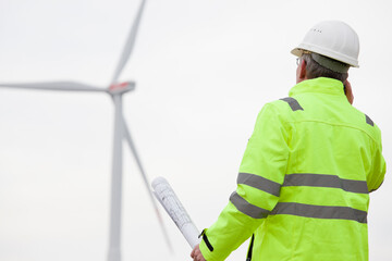 Mature engineer talking on mobile phone in front of a wind turbine with blueprint in his hand