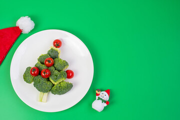 Christmas tree of organic vegetables on white plate; Broccolis and tomatoes with fork and knife on green background. Merry Xmas and happy New Year party, New You, Healthy food and vegetarian concept