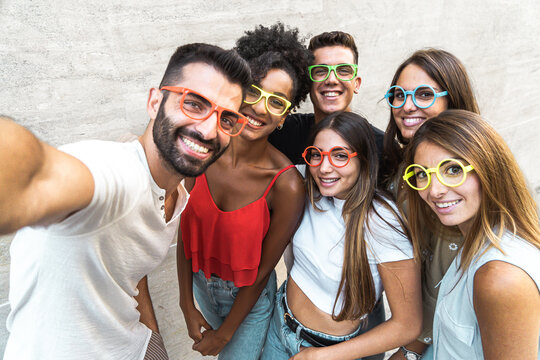 Millennial generation people group taking selfie with mobile phone wearing funny plastic glasses. Young people having fun together taking picture lifestyle concept