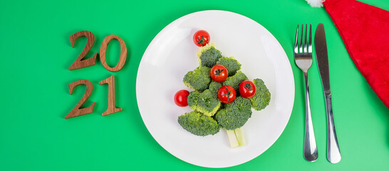 2021 Christmas tree of vegetables on white plate; Broccolis and tomatoes with fork and knife on green background. Merry Xmas and happy New Year party, New You, Healthy food and vegetarian concept