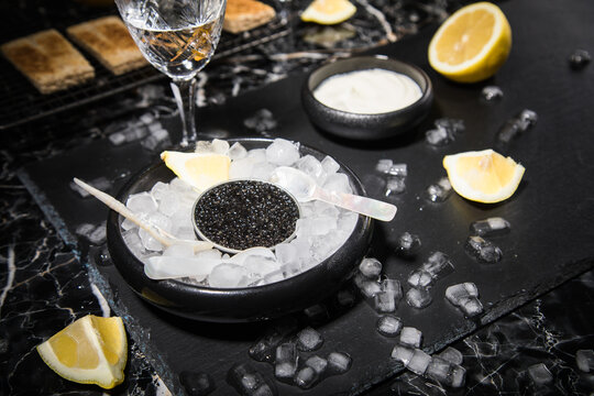 Schwarzer St&ouml;rkaviar auf Eis mit Perlmutt L&ouml;ffel, Schmand, Toast Brot, Zitrone und Wodka in Bleikristall Glas auf Schieferplatte und dunkel Marmor Hintergrund