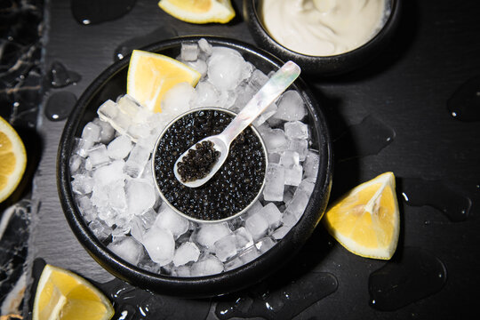 Schwarzer Störkaviar auf Eis mit Perlmutt Löffel, Schmand, Zitrone auf Schieferplatte und dunkel Marmor Hintergrund