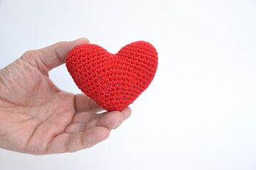 A man's hand holds between the thumb and forefinger a heart knitted of red yarn