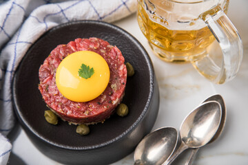Gourmet Rindertatar Beef Tatar roh aus Filet fettarm, Kapern, Eigelb, Zwiebeln auf Toast Brot und schwarz Teller mit Bier, Löffel, Geschirrtuch und Marmor Hintergrund hell