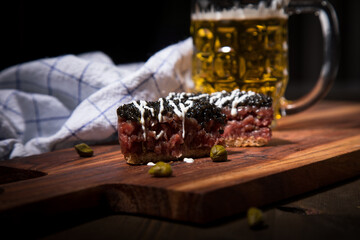 Gourmet Rindertatar Beef Tatar roh aus Filet fettarm, Kapern, Eigelb, Zwiebeln auf Toast Brot und schwarzer Kaviar, Schmand mit Bier, Küchentuch und Holzbrett Hintergrund dunkel