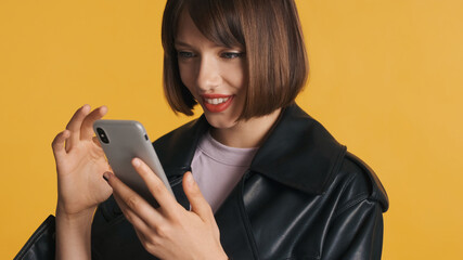 Naklejka premium Portrait of pretty girl with bob hair in casual wear happily surfes social media on smartphone isolated on yellow background