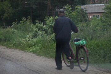 Russian elderly country man carries an old road bike on the side of an asphalt rural road on a summer evening against the background of green trees, village transport