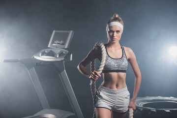 Sportswoman holding battle rope near treadmill and tire in gym with smoke
