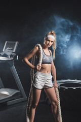 Selective focus of sportswoman with battle rope looking at camera near tire and treadmill in gym with smoke