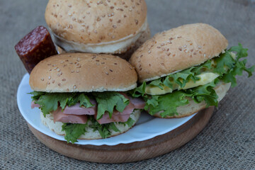 On a round board is a plate with two sausage, cheese and salad sandwiches. Buns and a piece of sausage lie nearby..