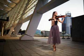 A beautiful adult woman with long hair posing with violin on pointe shoes outdoors in city.