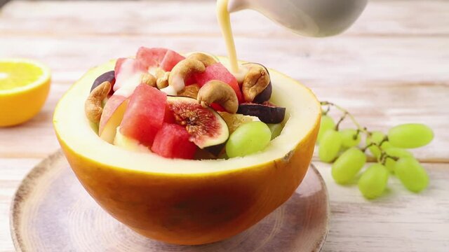 Vegetarian fruit salad of watermelon, grapes, figs, pear, orange poured with yogurt on white wooden background. Side view, close up.