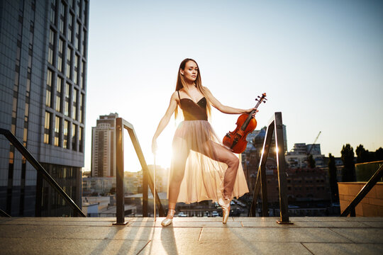 A beautiful adult woman with long hair posing with violin on pointe shoes outdoors in city.