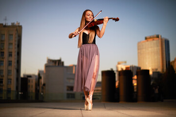 A beautiful adult woman with long hair posing with violin on pointe shoes outdoors in city.