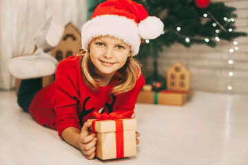 Obraz na płótnie Canvas Portrait of a pretty blonde girl in a Santa hat, lying on the floor with a gift in her hands on the background of a decorated Christmas tree. The concept of Christmas and New year. Holiday, gift