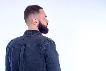 Fototapeta premium Portrait of a bearded man on a white background. The person is standing with his back to the camera and his head is turned to the side. Male portrait in profile. The guy in the denim jacket.