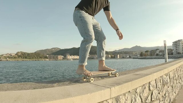 Young boy wearing blue jeans skating barefoot on the pier with the sea in the background