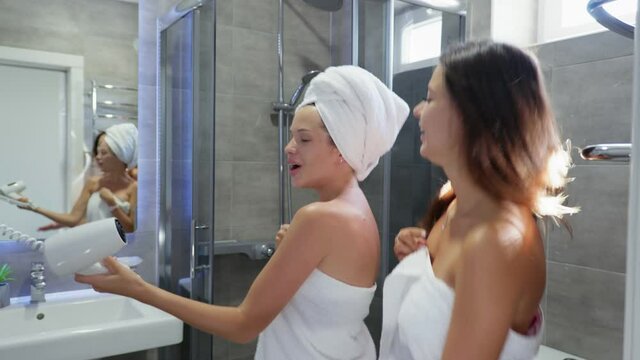 best friends, young attractive girls with beautiful skin in towel enjoy fun singing into hair dryer like microphone in bathroom