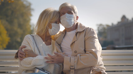 Senior couple with face mask embracing and holding hands sitting on park bench