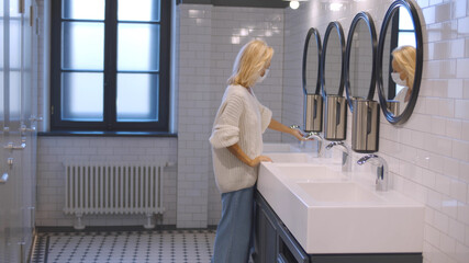 Mature woman in mask washing hands in public toilet