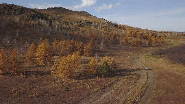 Aerial mountain off road trip. The SUV car quickly drives through the desert area along the mountains and autumn forest, leaving behind a raised dust and a long trail in the orange sand.