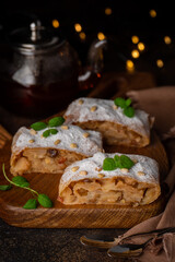 Homemade apple strudel, filled with apples, raisins, cinnamon and pine nuts. Traditional South Tirol recipe. Tea pot on background. Vertical orientation. Dark background.