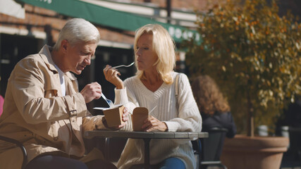 Aged couple sitting at small outdoor cafe table and enjoying takeaway lunch
