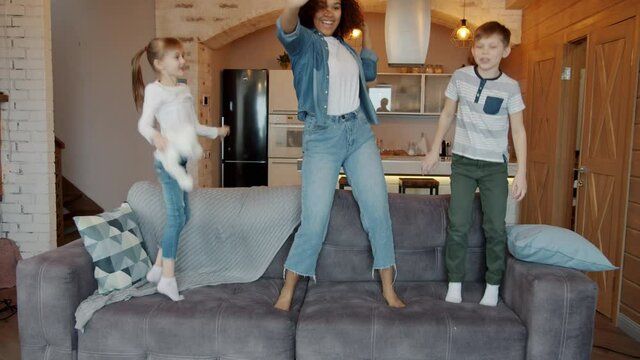 Playful woman babysitter and happy kids dancing jumping on sofa in apartment having fun together in leisure time. Youth and lifestyle concept.