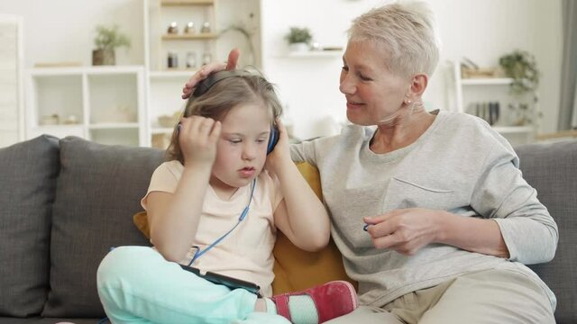 Lockdown of cute Caucasian girl with down syndrome wearing earphones sitting on sofa at home and listening to music while her good-looking grandmother sitting nearby and watching her